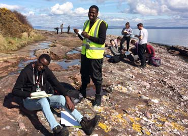two students holding notebooks, one sitting and one standing on an beach rock outcrop in arran