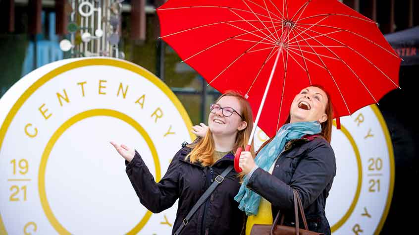 2/10/2021 - 2 women under a red umbrella laugh in the rain at the ChangeMakers Centenary Fesitval