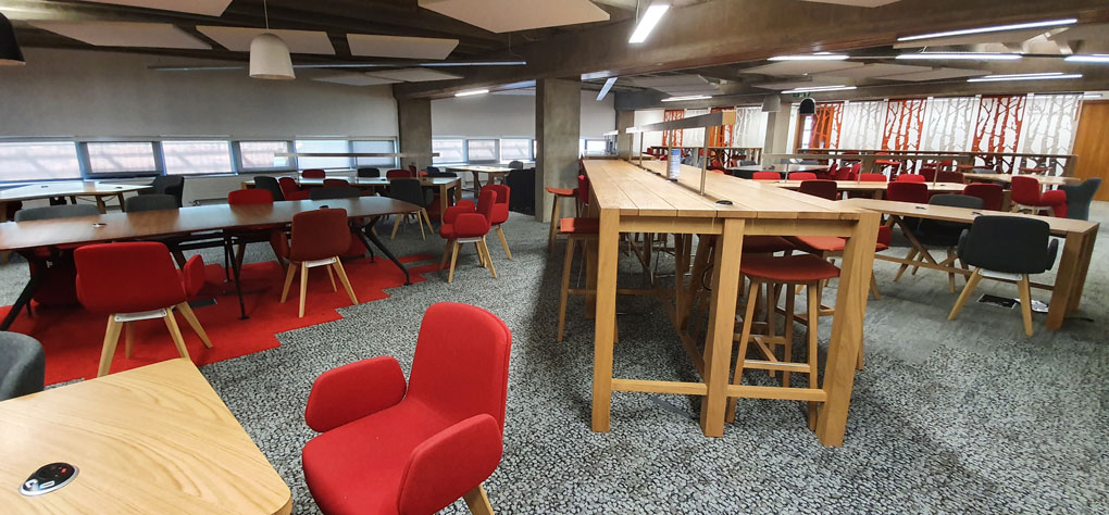 Various furnishing, red chairs and wooden tables of different heights, the carpet is red and there are hanging sound baffles on the ceiling. 