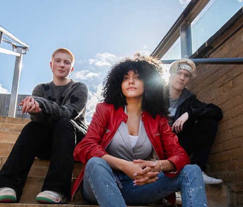 Three young people look down at camera while sitting on steps with buildings, bright blue sky and sunlight as backdrop