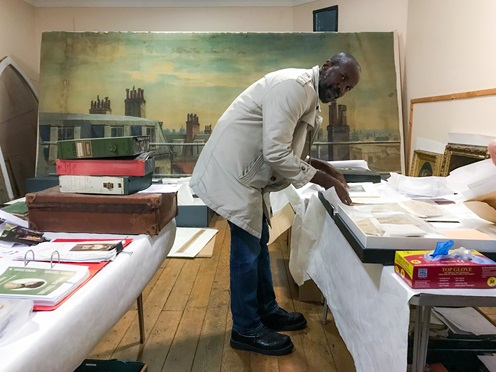 Man (David McAlmont) wearing a white coat, leaning on a table with various archival objects and books. Books are inside boxes with several sheets of tissue paper. In the background an old painting of a skyline (roofs and chimneys partially visible)..