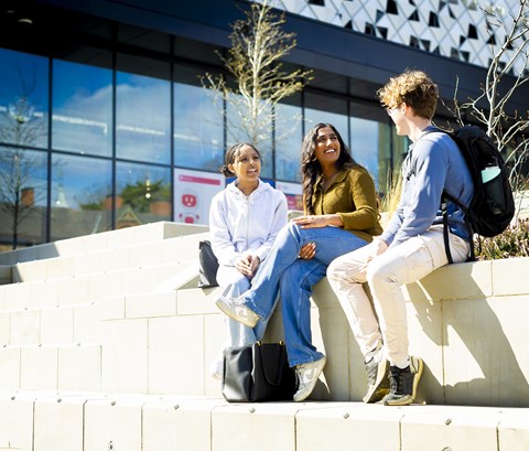 3 students smiling and chatting, sitting in the sunshine, outside Freemen's Bar & Kitchen in the Student Village