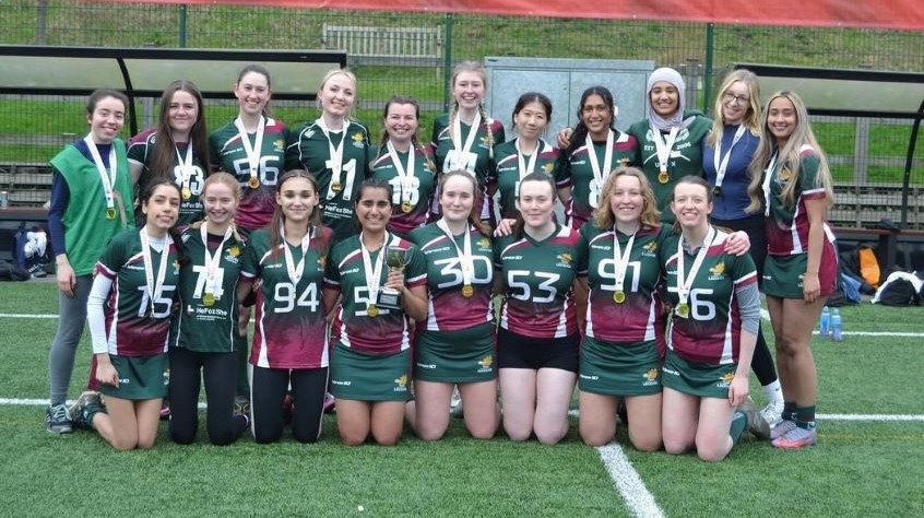 An image of the women's lacrosse team, smiling at the camera, each with a medal around their neck to celebrate their Varsity win