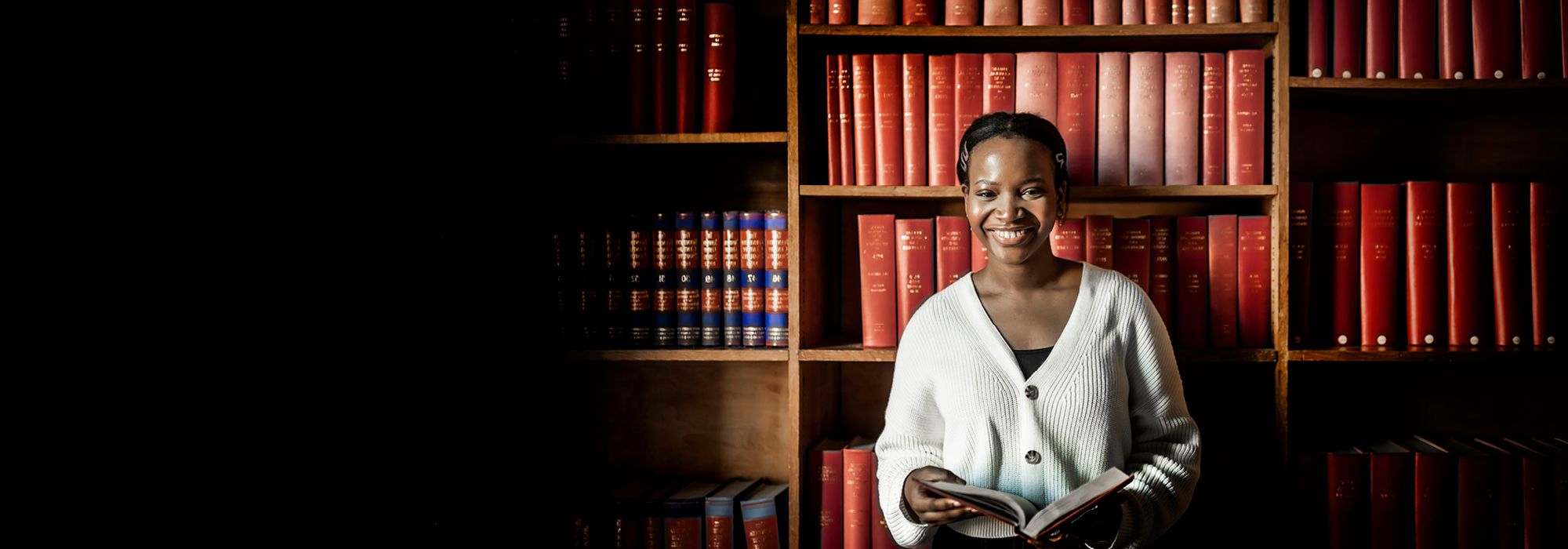 A person stands in front of tall wooden bookshelves filled with red and dark-bound volumes, holding an open book. Warm lighting illuminates the subject and surrounding shelves.