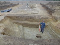 View of the defensive ditch at Ebbsfleet during the 2016 excavation, a stepped ditch with a square base. A man stands in the ditch holding equipment to illustrate its depth, which is above his head