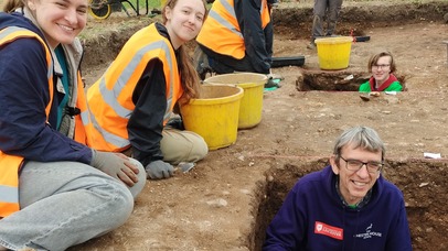 Two students in high vis jackets sitting on the dirt, member of staff standing shoulder-deep in a trench