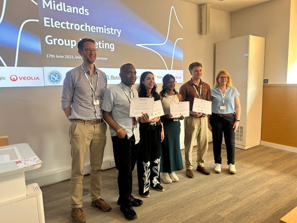 Prize winners at the Midlands Electrochemistry Group meeting. Dr Andy Wain, and Dr Emma Smith, presenting prizes to the winning students at the conference, standing in a lecture theatre with a backdrop of the meeting logo - a cyclic voltammogram on a glossy blue background.