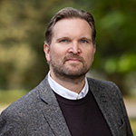Photograph of Gustav Berggren, wearing a suit and standing outside in front of a background of trees.