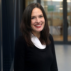 Photograph of Serena DeBeer standing in a corridor, smiling.