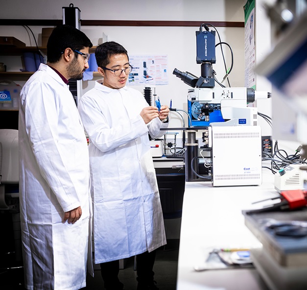 Jake Yang teaching a student how to use equipment in a laboratory. The laboratory is white and clean, and Jake is standing in front of a microscope.