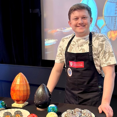 Josh Smalley presenting at the Edinburgh Science Festival. Josh is wearing a University of Leicester apron, standing behind a table covered in chocolate eggs, bakes, and biscuits in the shape of rockets.