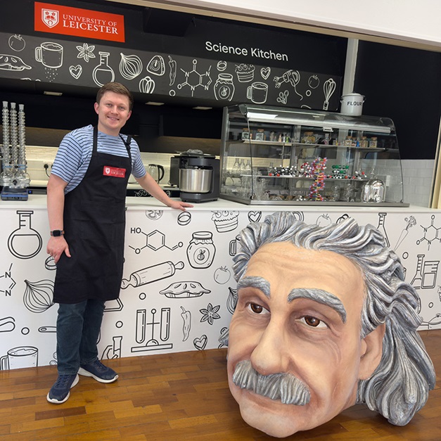 Dr Josh Smalley, wearing a University of Leicester apron, standing in the Science Kitchen. The room contains a large cartoon bust of Einstein and counter for serving and displaying scientific instruments and cakes.