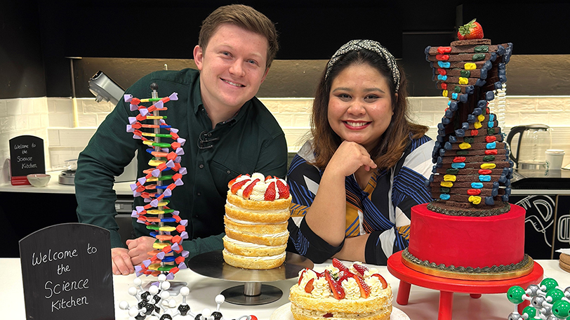 Josh Smalley and Syabira Yusoff from the Great British Bake Off standing behind models of DNA, and a cake in the shape of a DNA double helix and other baked goods.