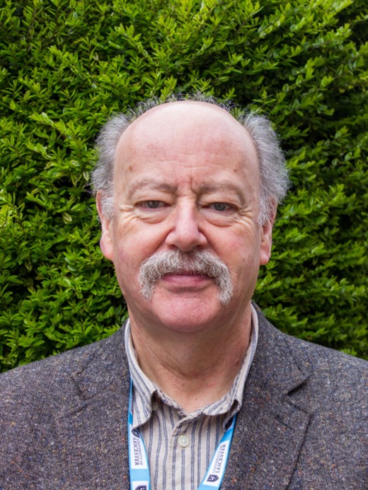 A photograph of Robert Hillman. An older white male, with a large moustache. He is wearing a grey suit jacket over a striped shirt. He is standing in front of a green tree.