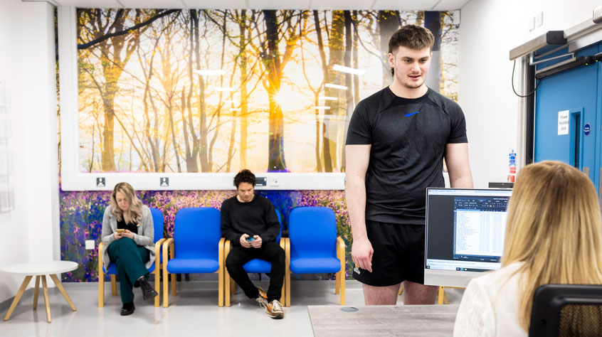 A patient approaching a receptionist whilst other patients wait in the background.