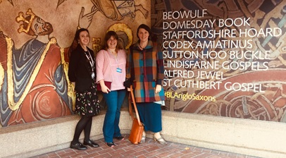 Group of three women posing for a photograph in front of a large reproduction of a medieval artwork advertising the British Library Anglo-Saxon Kingdoms exhibition