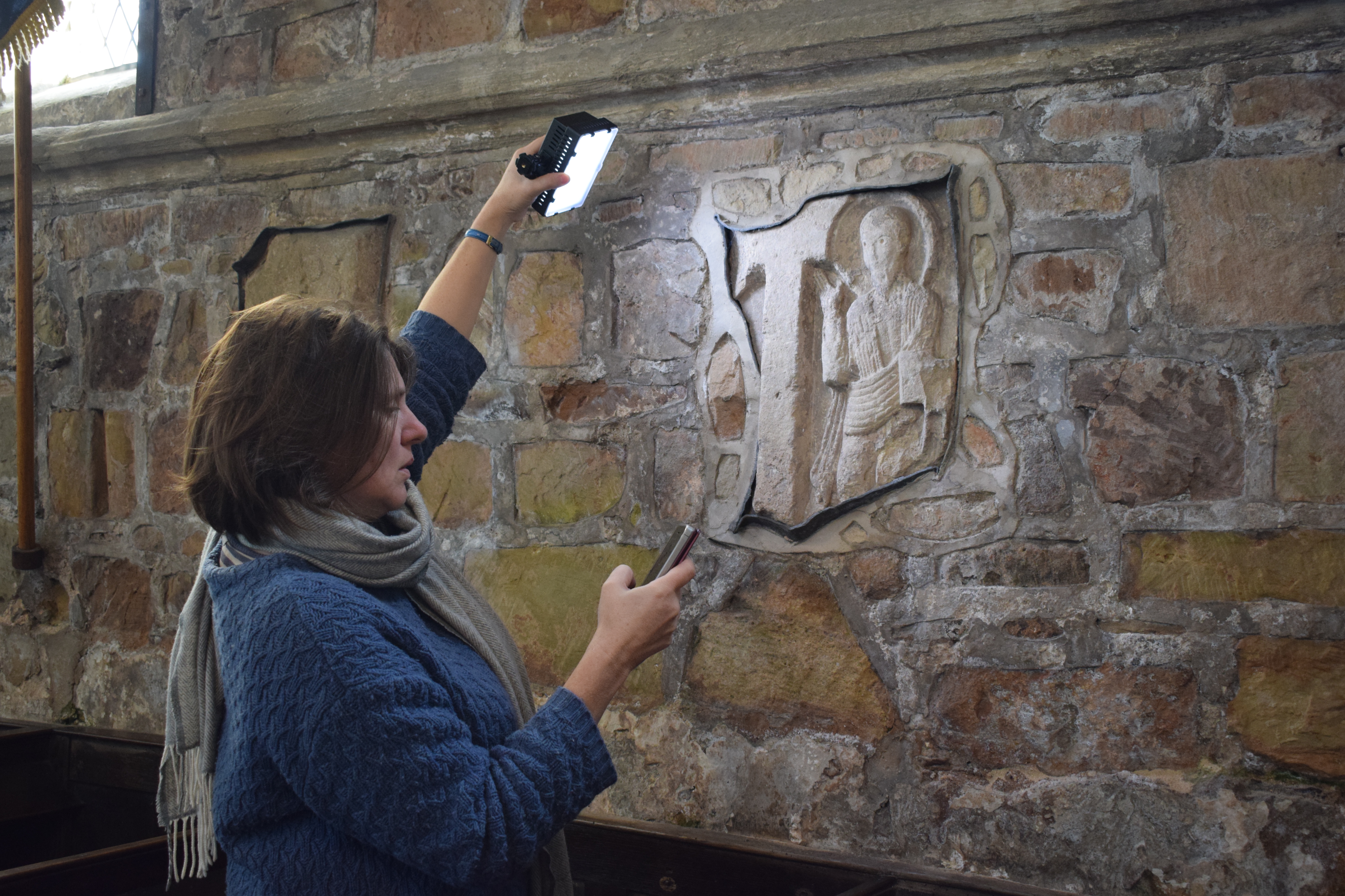 Woman with medium length hair and wearing a blue jumper and scarf holding up a bright light and another device to study a depiction of a figure sculpted into a stone wall