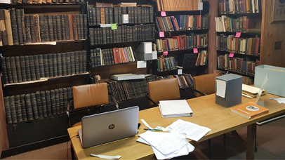 Desk with laptop and notes, with archive book shelves behind