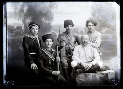 A group of five men posing for a photograph. One is holding a gun, another has ammunition on a belt across his torso. Three of the men are wearing hats.