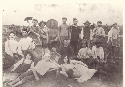 A group of people posing for a photograph - some standing, others sitting or laying on the ground - on a farm. Many are holding farming tools.