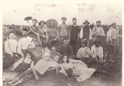 The Oded Yarkoni Historical Archives of Petakh Tikva, Moshav Ein Ganim, around 1908, Sign. 002-002/2514 A group of people posing for a photograph - some standing, others sitting or laying on the ground - on a farm. Many are holding farming tools.