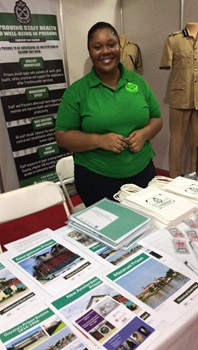 Project pamphlets on display at the Guyana Prison Service Annual Service of Thanksgiving, 23 September 2022. Behind the table of pamphlets is a smiling woman wearing a green Prisons Service t-shirt and some khaki uniforms on mannequins.