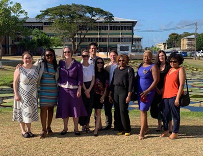 The project team, University of Guyana, April 2019. A group of 11 people pose for a photograph in front of a pond. In the background are some trees and large office-like buildings.