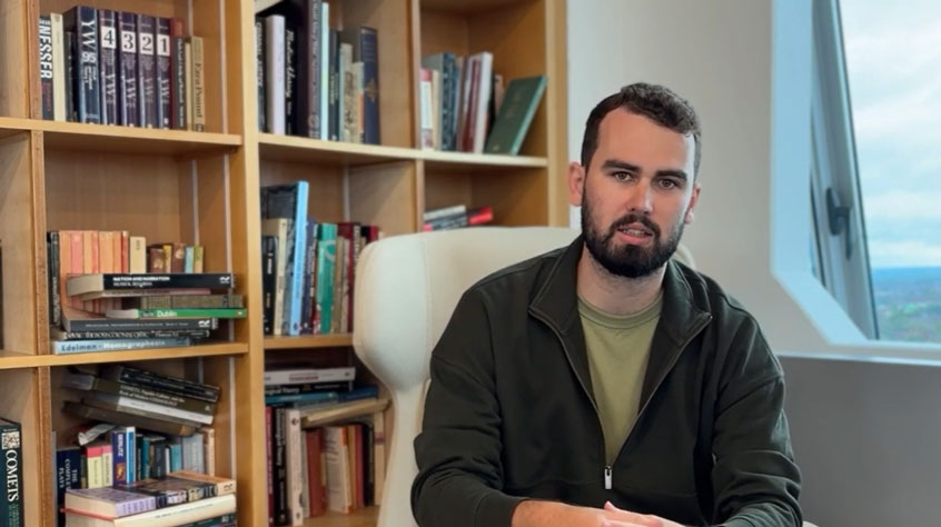 Man sitting in front of a bookshelf, talking to the camera.