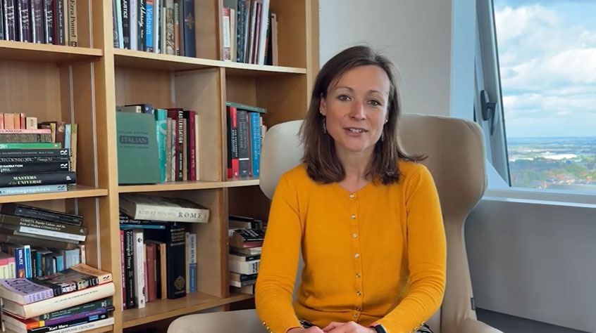 Woman sitting in front of a bookshelf, talking to camera.