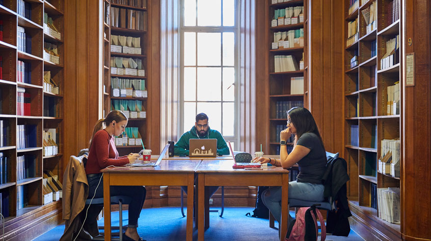 Three students sitting at a table in an ornate library, with large shelves of books around them