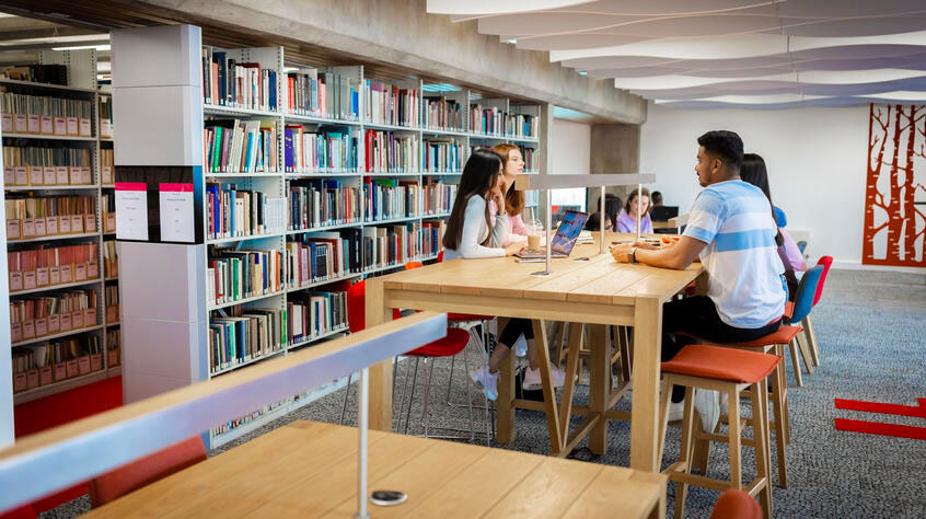 Students sitting at a high table in a library with a large shelf of books behind them.