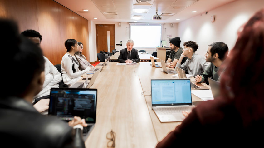 Students and a lecturer sitting around a large conference table, some with laptops open in front of them.