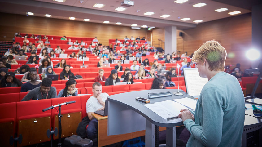 A large lecture hall with red bench seats. Over half of the room is full of students.