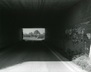 Black and white photograph of a road under a tunnel with a sharp turn chevroned sign at the far end