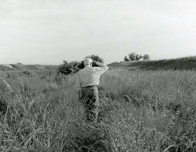 Black and white photograph of an older man in the middle of a grassy meadow, grasses up to his waist, looking away from the camera into the distance