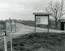 Black and white photograph of a road with a park map station in the centre-right of the picture on a grass half of ground