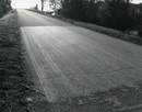 Black and white photograph of a wide paved road, a crack across its width in the foreground  as sunlight catches a difference in tarmac colour in the centre