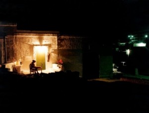 Photograph taken at night, mostly dark except for a man sitting outside a stone building with an outside light illuminating the area above and around him