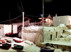 Photograph of a stone dock at night with small fishing boats on land in the foreground and a lit building in the background