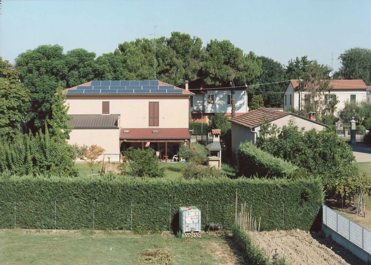 Photograph of a light pink house surrounded by a green hedge with a plot of land in the foreground and other houses nearby