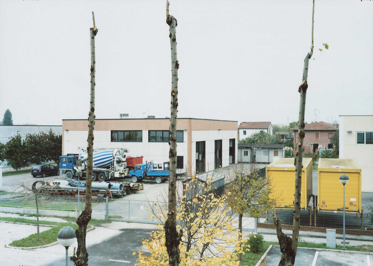 A photograph at a high angle of a car park behind a depot containing a cement mixing truck and other vehicles, with three slim tree trunks mostly stripped of branches in the foreground