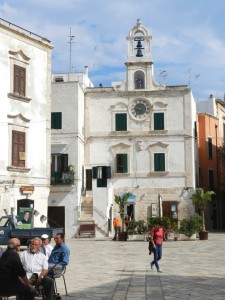 Photograph of a town square with people sitting and walking around and building with a clock tower in the background