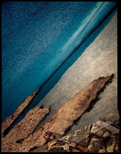 Photographic artwork based on coastal seascape featuring jagged rocks, deep and dark colour contrast between ocean, beach, rocks and stone wall