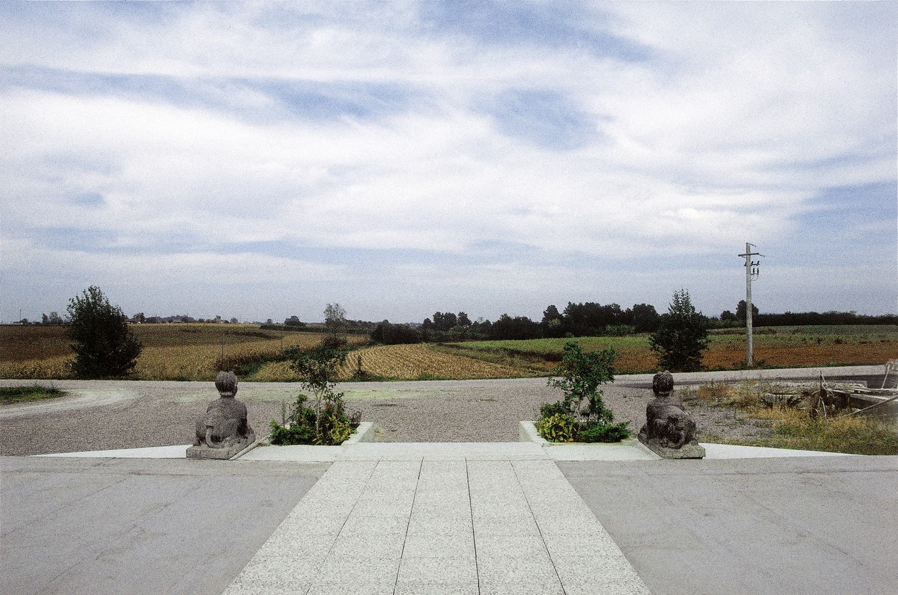 Photograph taken from a raised concrete patio, sitting animal statues either side of some stairs looking out to fields
