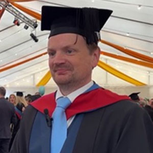 Man wearing a graduation cap and gown and standing in a marquee.