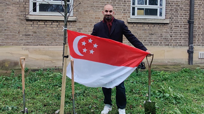 Person holding a flag of Singapore, whilst planting a tree on University of Leicester campus.