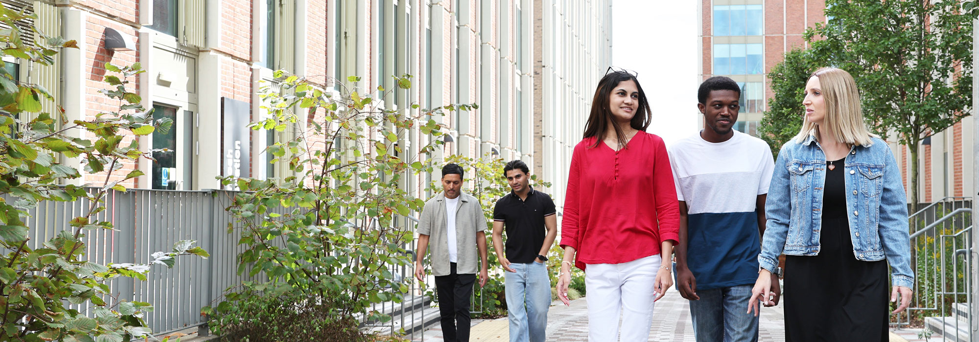 A group of university students walk along a landscaped pedestrian pathway between modern campus buildings, with trees and greenery lining the route.