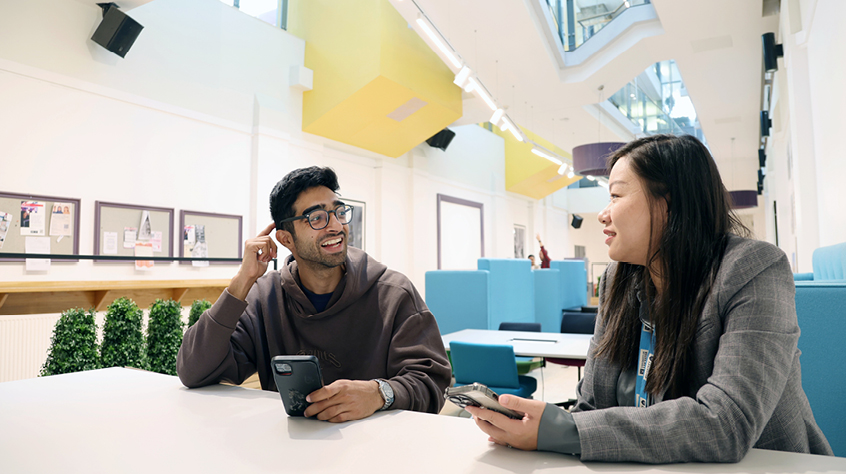 Two people using mobile devices while talking at a table in a bright university learning space.
