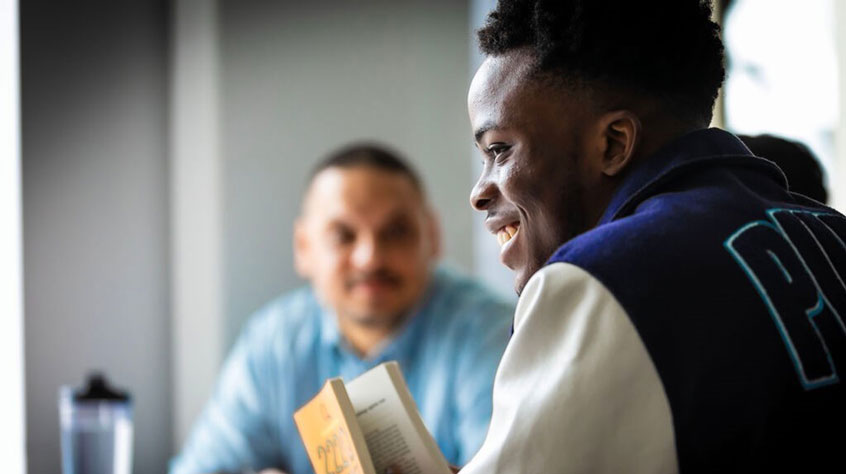 Student sitting at a table, holding a book and smiling.