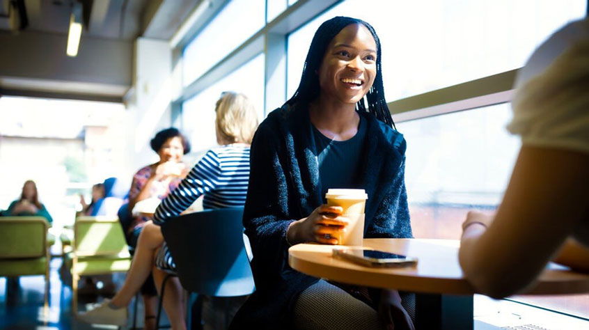 Student sitting at a table in the library cafe, holding a coffee and smiling.